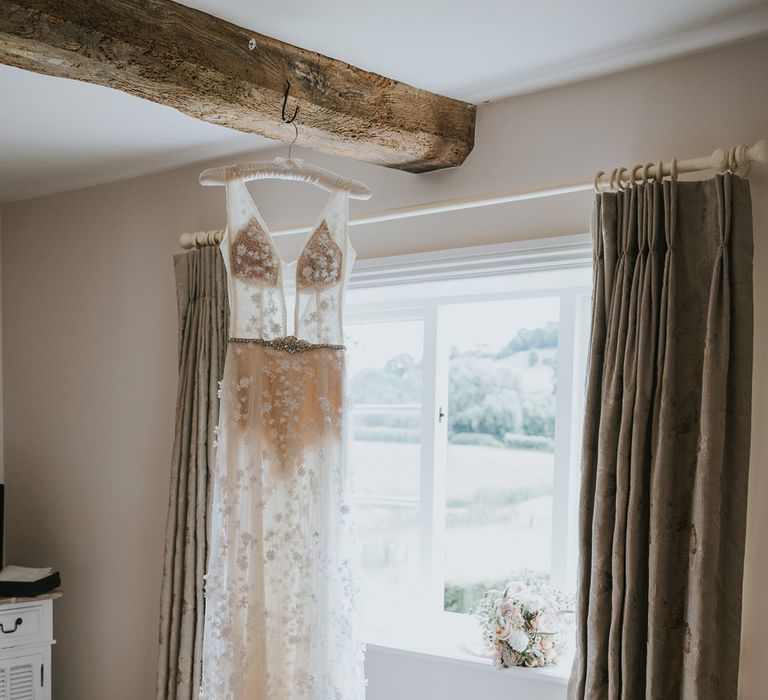 Sheer applique Berta wedding dress hanging from wooden beam by window with strap heels on the floor at Primrose Hill Farm wedding