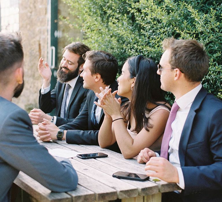 Wedding guests sitting on benches at outdoor drinks reception 