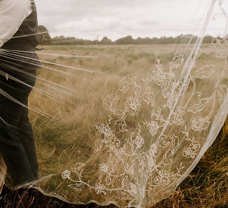 Brides veil blows in the wind whilst stood in a field complete with fine lace florals to the bottom of train