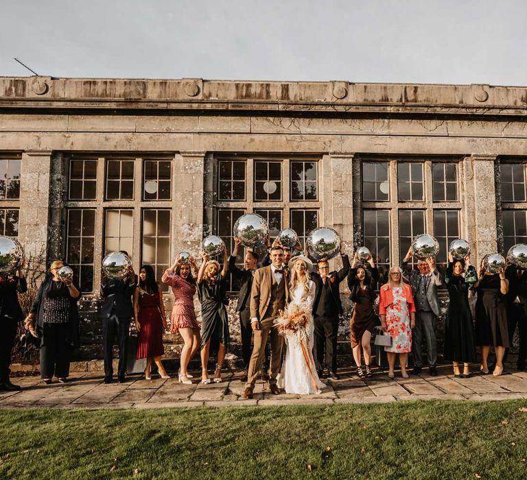 Bride in hat and boho wedding outfit holding white and orange bridal bouquet stands with groom in three piece brown tweed suit and wedding guests all holding disco balls outside Kinmount House in Scotland