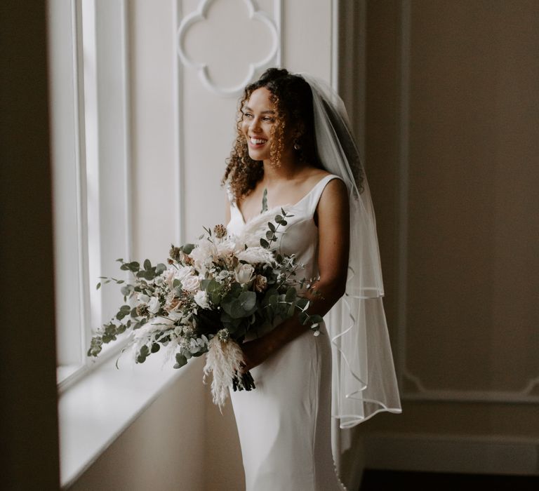 Bride looks out the window on her wedding day whilst holding floral bouquet