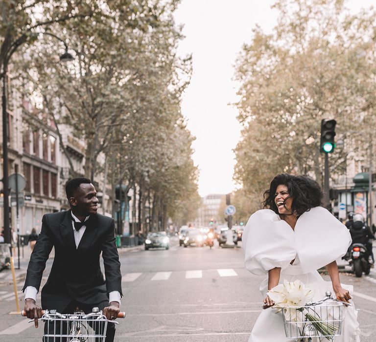 Black groom in a tuxedo riding a bicycle through Paris with his South Asian bride in a puff sleeve wedding dress with plunging neckline