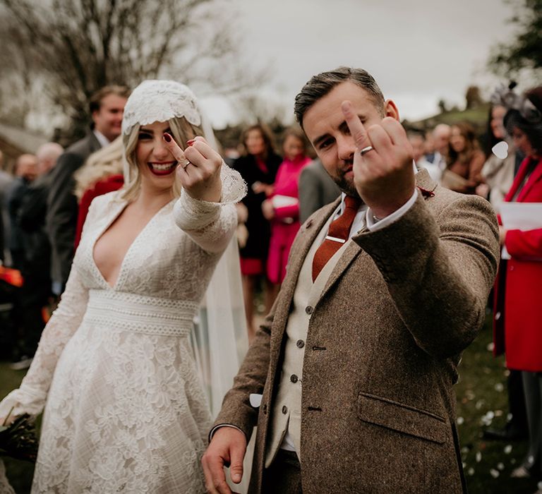 Bride in a lace wedding dress and Juliet cap veil and groom in a wool suit holding up their ring fingers 