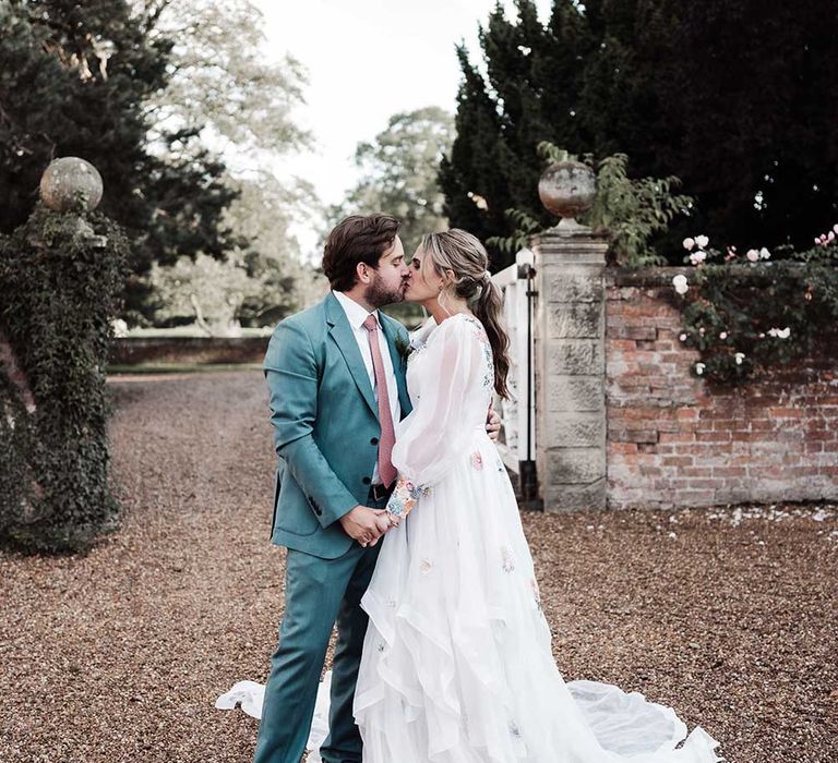 Groom in blue suit and pink tie kisses bride in long sleeve Victoria Sanders wedding dress with embroidery and train in the grounds of Iscoyd Park