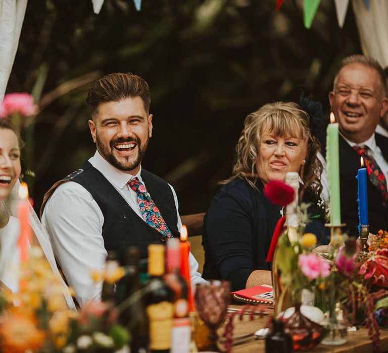 Groom in a navy blue waistcoat and floral Liberty print tie laughing during the clear marquee wedding reception speeches 