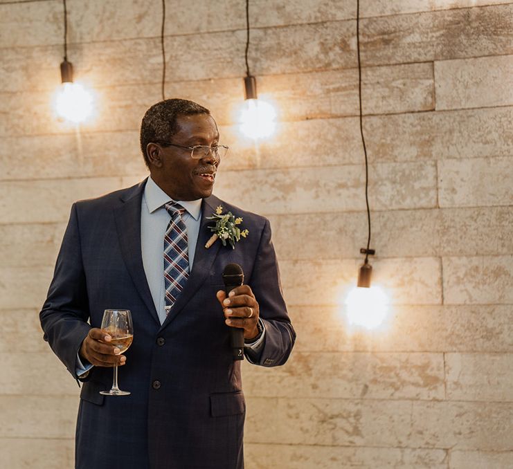 Father of bride stands with glass of wine in hand in fitted suit with stunning boutonniere as he gives a speech