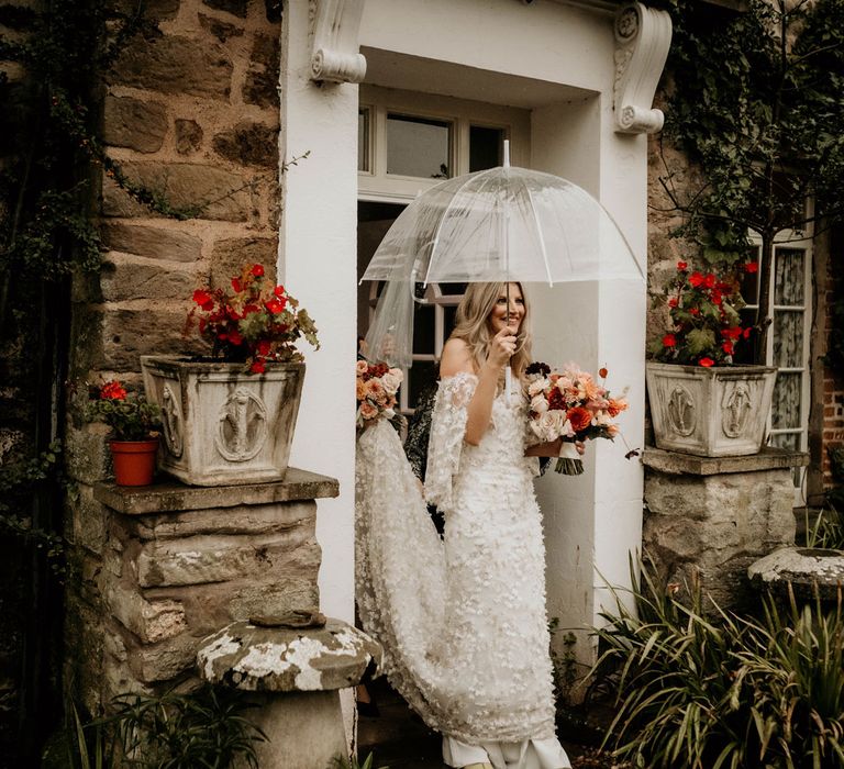 Bride in applique wedding dress with 3d flowers holding mixed dusky bridal bouquet and clear umbrella walks out of stone house before wedding ceremony