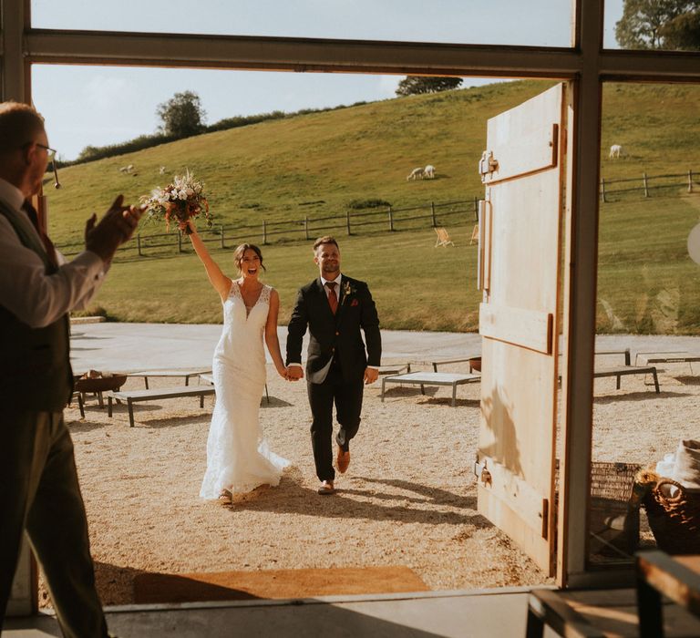 Bride in v neck lace wedding dress with train holds up bouquet as she walks in reception holding hands with groom in dark suit with burgundy tie