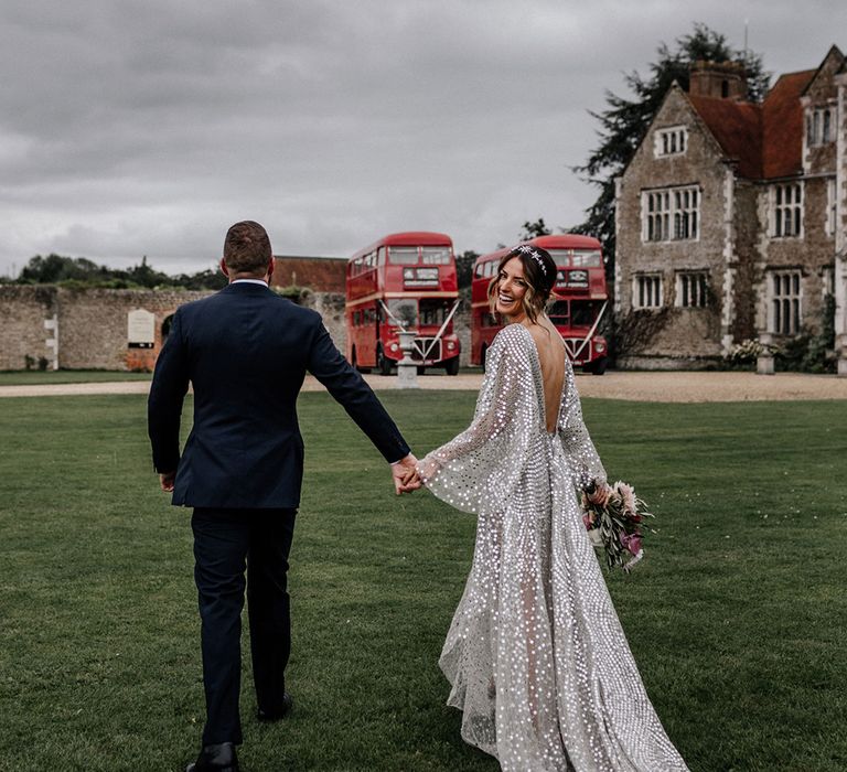 Groom in navy blue suit holding hands with his bride in a silver disc wedding dress with loose sleeves and low back detail 