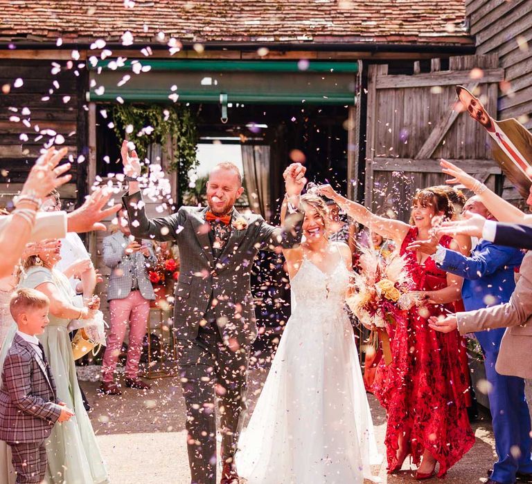 Bride in lace wedding dress with train holding mixed dried floral boho bouquet holds up arm with groom in green three piece suit and pattered shirt as guests throw petal confetti