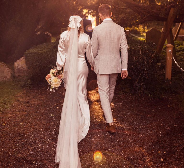 Bride in satin hair bow and veil walks holding hands with groom in light grey suit through churchyard after wedding ceremony 