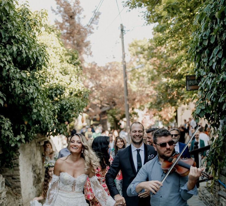 Bride & groom walk with wedding guests as violinist plays in front of them as they walk 