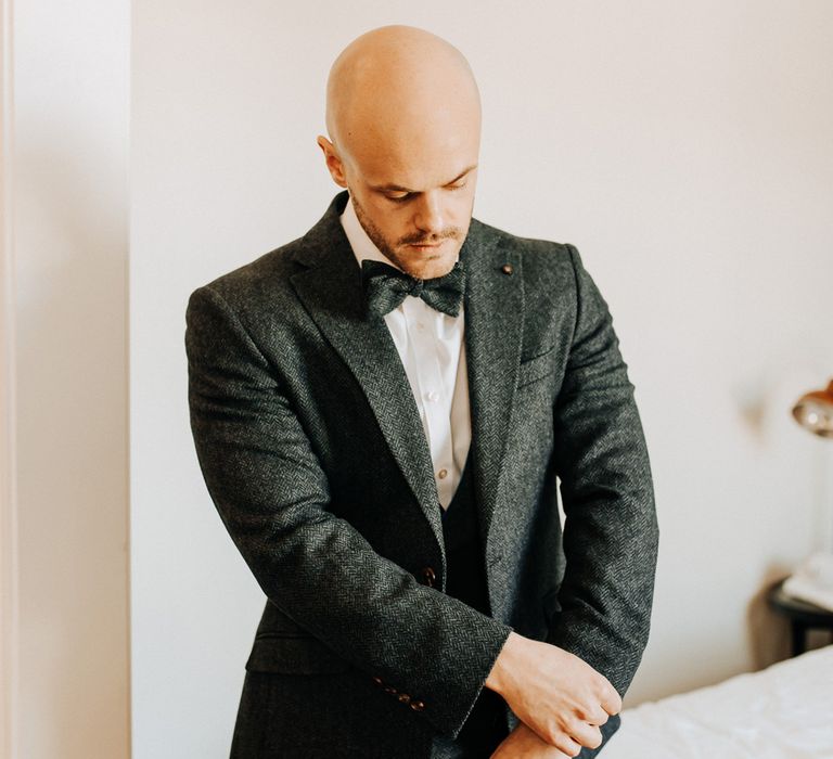 Groom in dark three piece tweed suit with matching bow tie adjusts cuffs as he gets ready for wedding