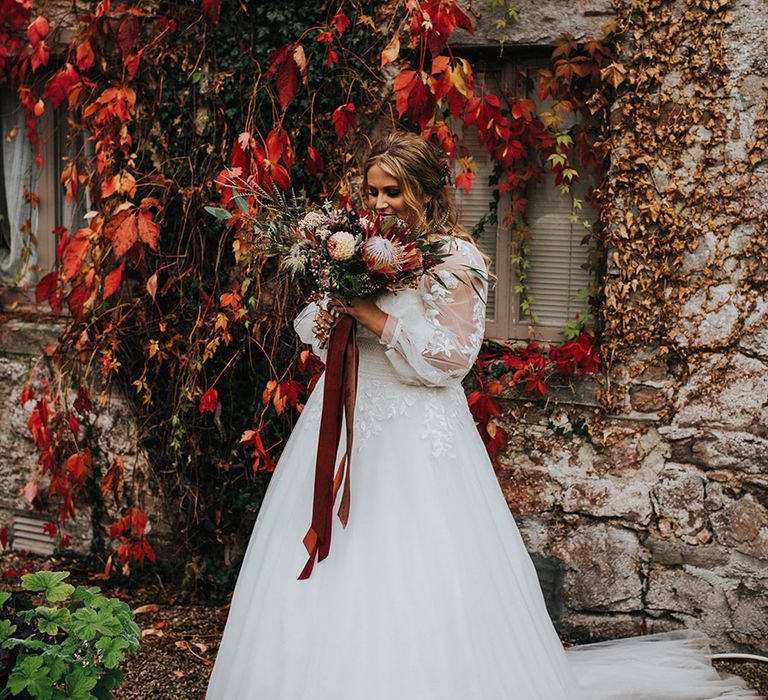 autumn theme wedding at Askham Hall bride in a princess wedding dress with long sleeves looking at her protea wedding bouquet tied with ribbon 