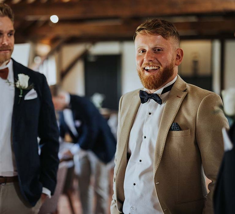 Groom in beige suit jacket and groomsman in dark suit jacket ad burnt orange coloured bow tie for rustic wedding