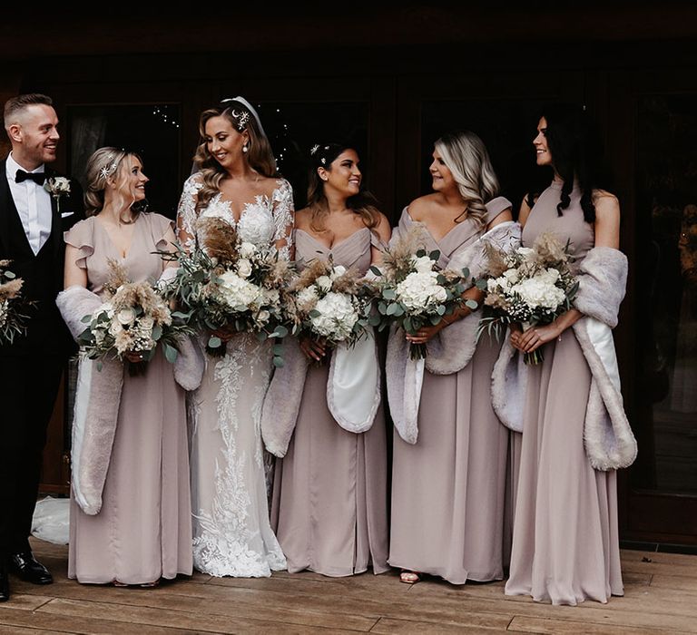 Bride in lace wedding dress standing with bridesmaids in taupe coloured dresses with various neckline styles and matching fur shawls