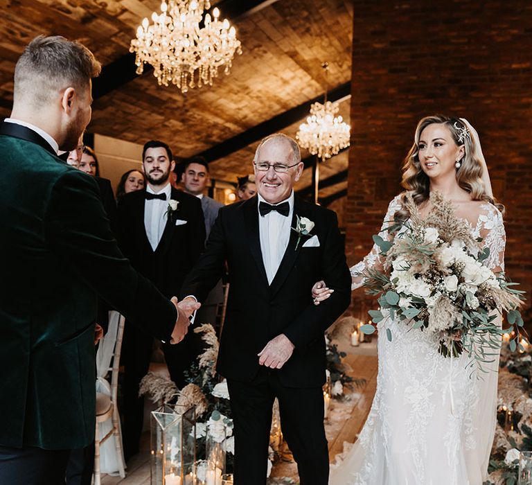 Father of the bride in black tie shakes hands with the groom in green velvet suit jacket as he walks daughter down the aisle