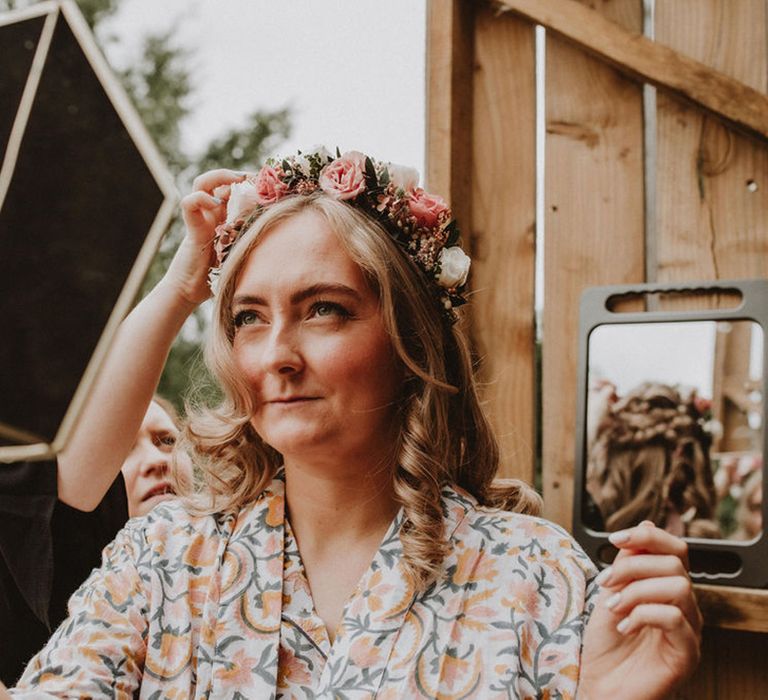 Bride holds up a mirror as she gets her hair done in an intricate braided medieval hairstyle with flower crown for wedding day