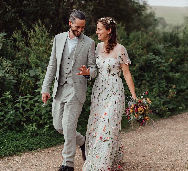 Groom in grey smiles at his wedding ring as he walks with the bride in floral wedding gown and flower hair accessories 