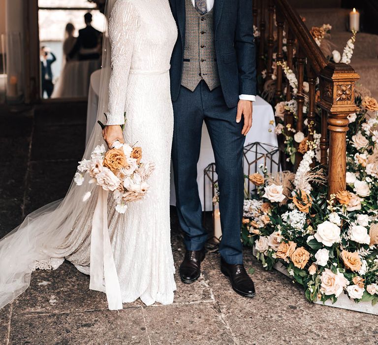 Groom in dark blue suit with patterned waistcoat and paisley tie poses with bride in beaded wedding dress next to floral display