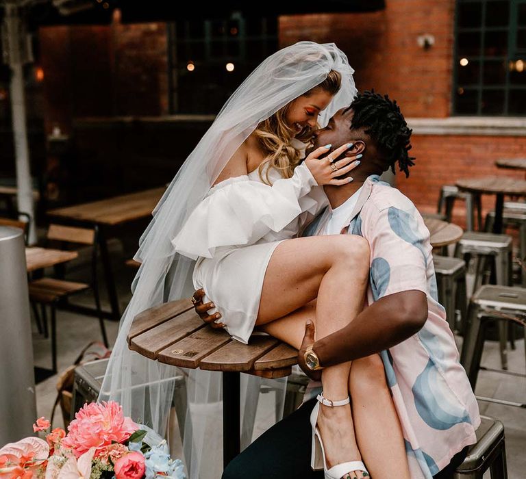 bride in a short wedding Dress and platform shoes sitting on a table with her groom in a patterned shirt embracing her