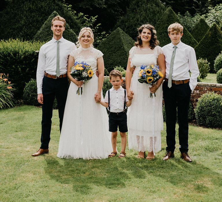 Brides in sheer high neck wedding dresses hold hands with young boy and hold wedding bouquets with sunflowers and gypsophila with older children next to them