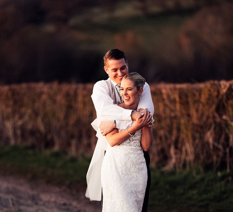 Groom wearing grey tie hugs the bride from behind wearing lace wedding dress