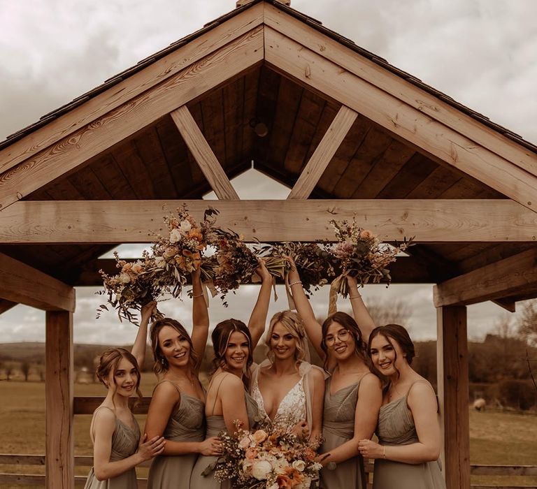 Bridesmaids in green dresses with the bride in lace wedding dress lifting orange and white bouquets 