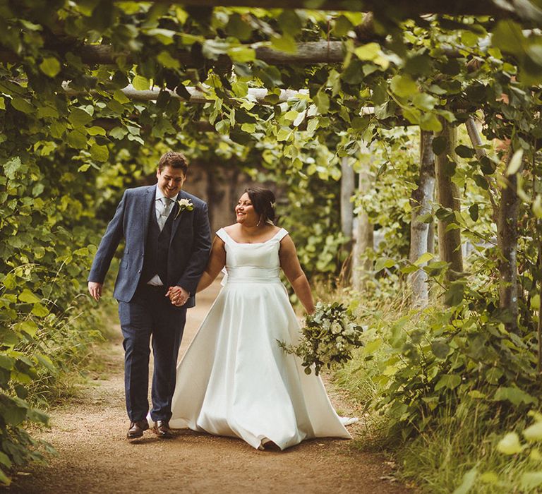 Bride in princess skirt wedding dress and groom in blue suit walk hand in hand in the gardens of Hatfield House