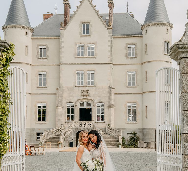 Bride leans against bride with blonde hair as they pose together in their lace wedding dresses