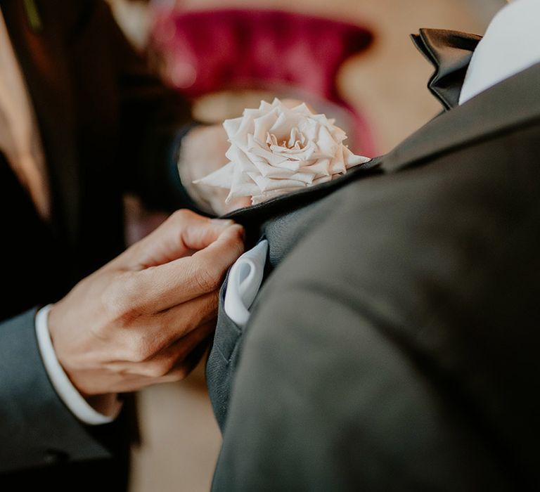 Groom gets help attaching his rose buttonhole to his tuxedo 