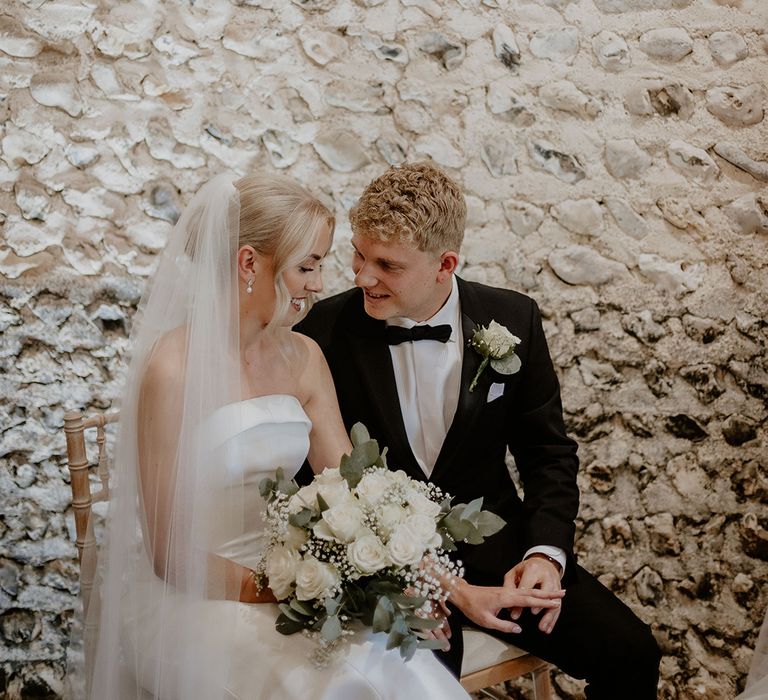 Groom in black tie with white rose buttonhole sits with bride in strapless gown with white rose bouquet during their ceremony 