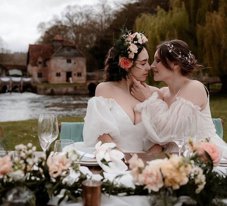 Bride in an ethereal wedding dress with sheer sleeves kissing her bride in a strapless wedding dress at their sweetheart table 