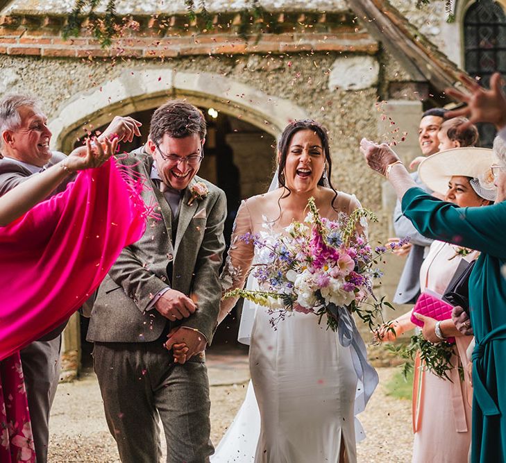 Bride wearing Hannah Elizabeth wedding dress with sheer embellished long sleeves holds colourful floral bouquet walking through confetti