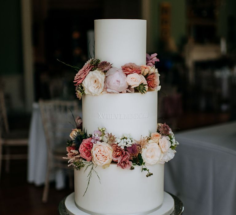 Three tier wedding cake complete with white frosting and embossed latin numerals alongside floral decoration