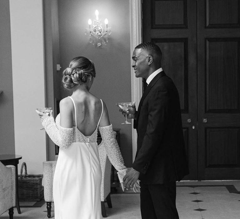 Bride in evening wedding dress with beads and off the shoulder detail walking with groom in black tie as they drink cocktails 