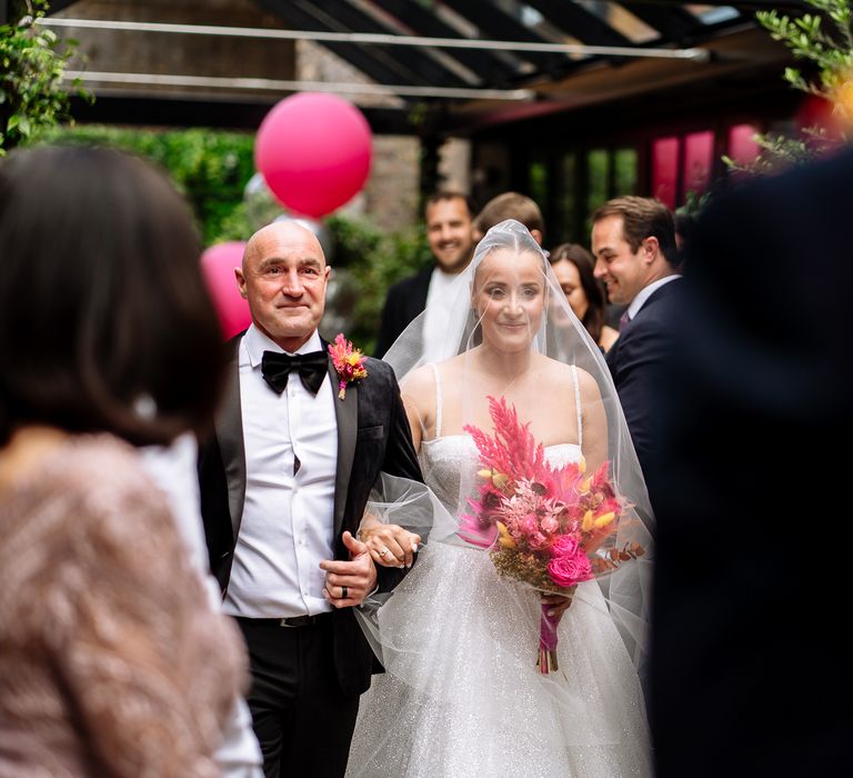 Bride holding colourful dried floral bouquet with pink pampas grass and with veil across her face walks down the aisle with her father