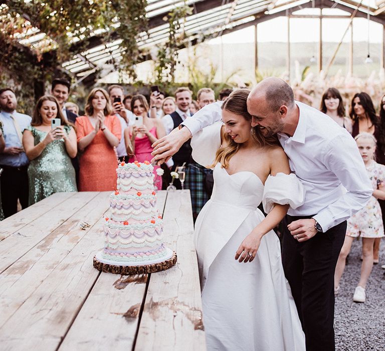 Three tier colourful pastel retro style wedding cake is cut by the bride and groom as the guests watch