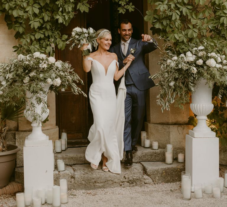 Bride holds the train of her Suzanne Neville wedding dress and enters the reception with her groom between two floral arrangements 