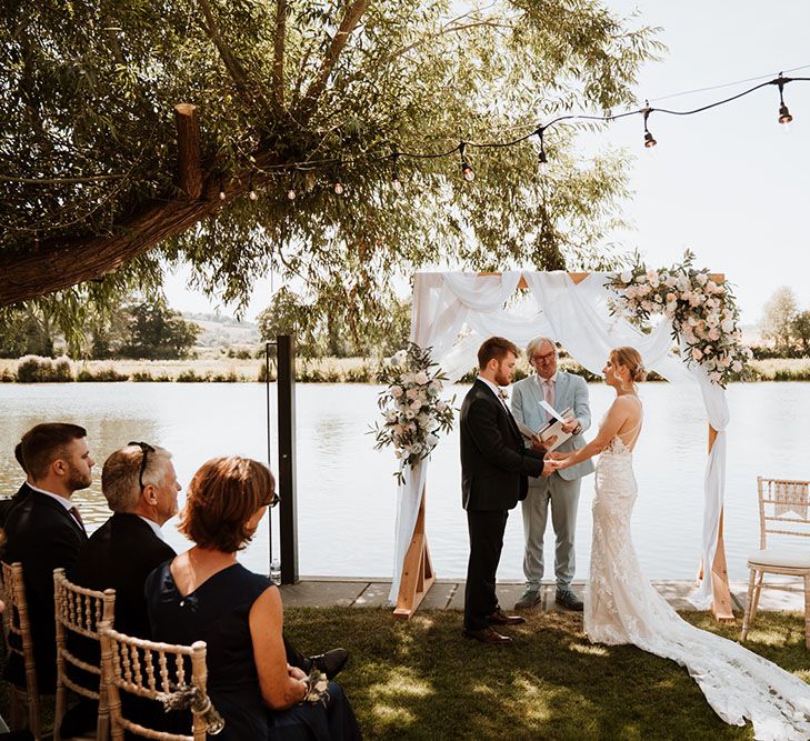 Bride & groom stand in front of wooden arch complete with white fabric draped across and floral decor 