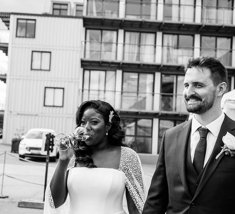 Bride sips champagne on her wedding day as she walks with her husband at Trinity Buoy Wharf 