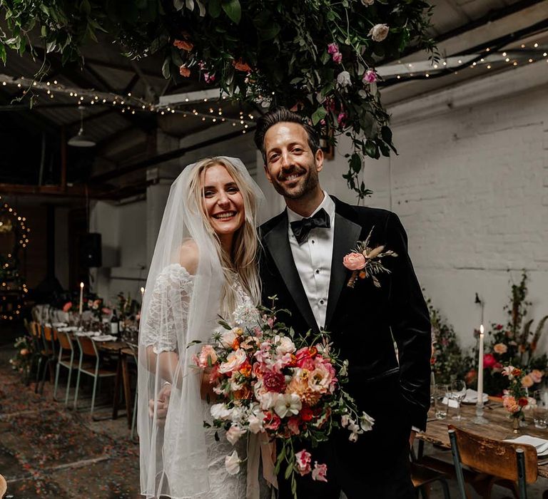 Bride in boho two piece lace bridal outfit with large colourful mixed bouquet and groom in black tux with velvet look bow tie