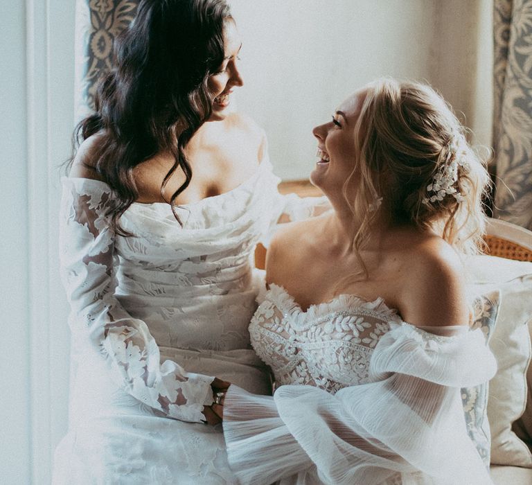 Bride in off the shoulder long sleeve wedding dress sitting on bride's lap wearing strapless lace wedding dress on cream coloured chair, laughing together