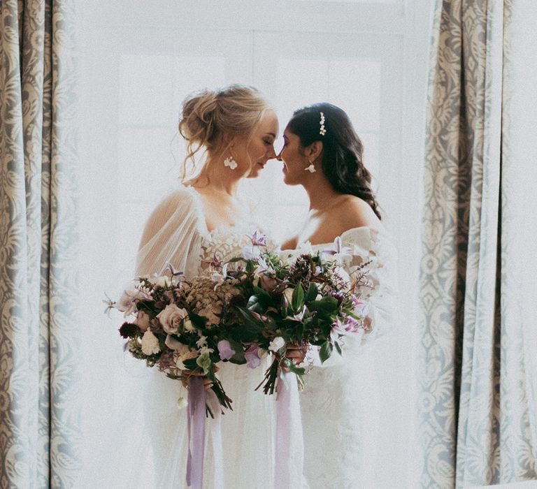 Brides in complimenting lace detail long sleeve wedding dresses holding matching bridal bouquets with ixed neutral floral bouquet with white carnations, purple tulips, roses and foliage