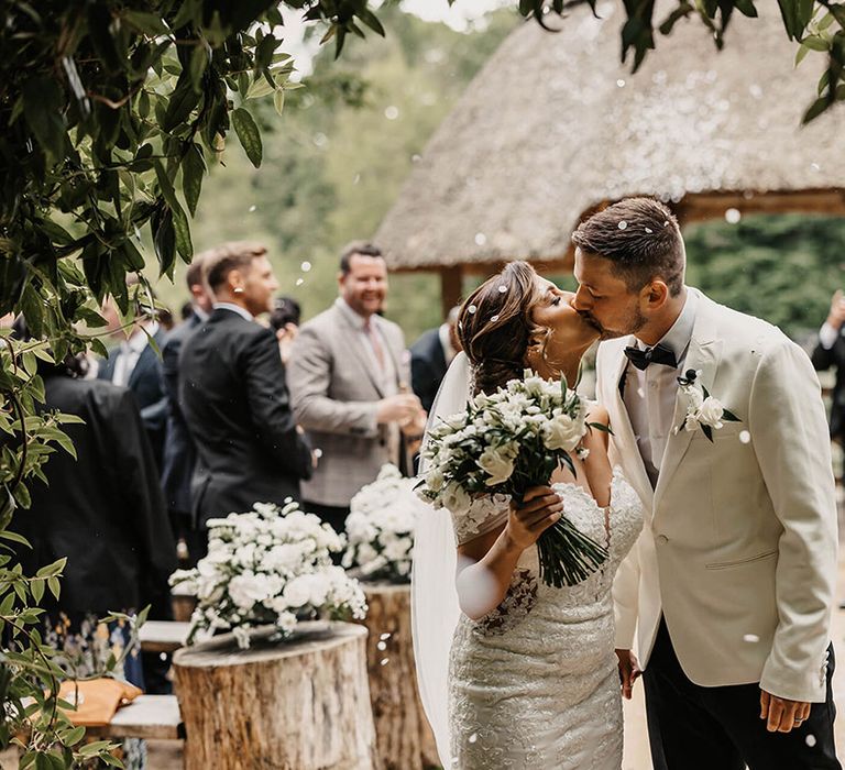 Bride in flower lace wedding dress kisses the groom in a white tux after their confetti exit 