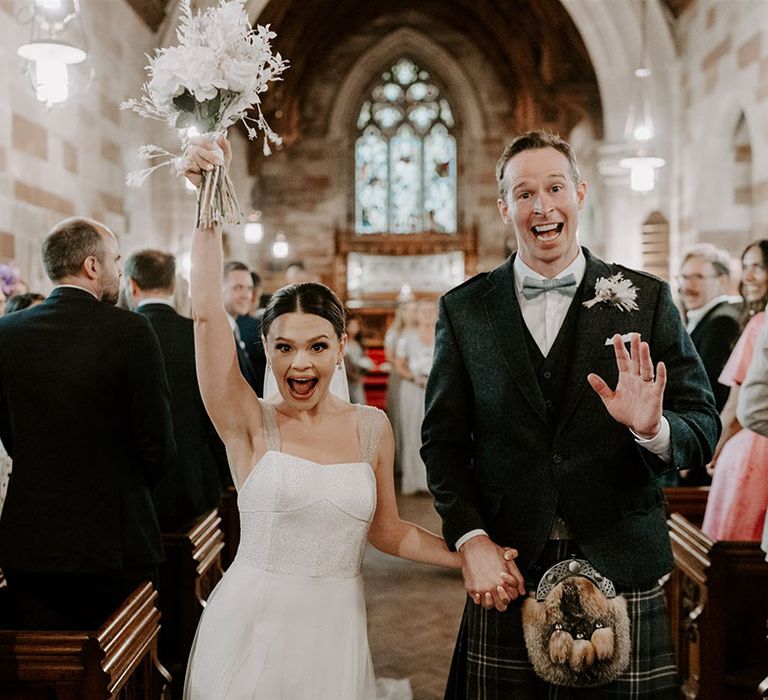 Ecstatic bride lifts up her white bouquet with dried flowers walks out of the church with the beaming groom in a kilt