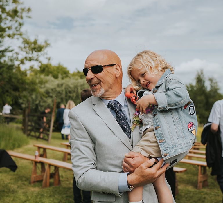 Wedding guest holds little boy in personalised denim jacket during outdoor wedding ceremony 