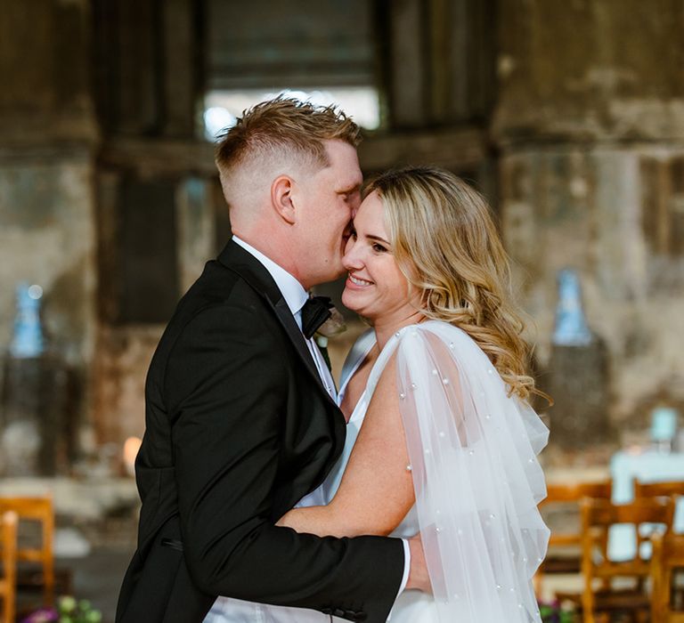 Groom wearing black tie holds his bride in pearl embellished wedding cape and side split wedding dress