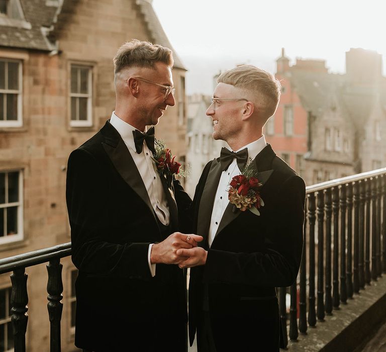 Grooms in matching black velvet tuxedos with black bowties and red rose and foliage boutonnieres holding hands and looking into each other's eyes on the rooftop of The Balmoral Edinburgh 