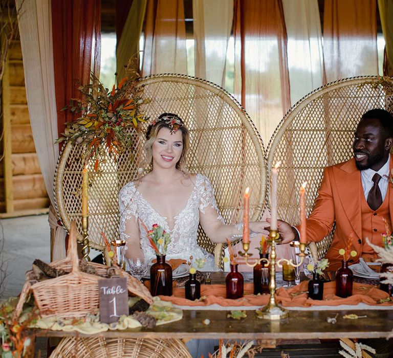 Bride and groom at autumnal harvest themed wedding tablescape with orange and yellow tapered candles, gold candlesticks, dark orange bottles with dried flowers, and naturally dyed cheesecloths for tablerunners